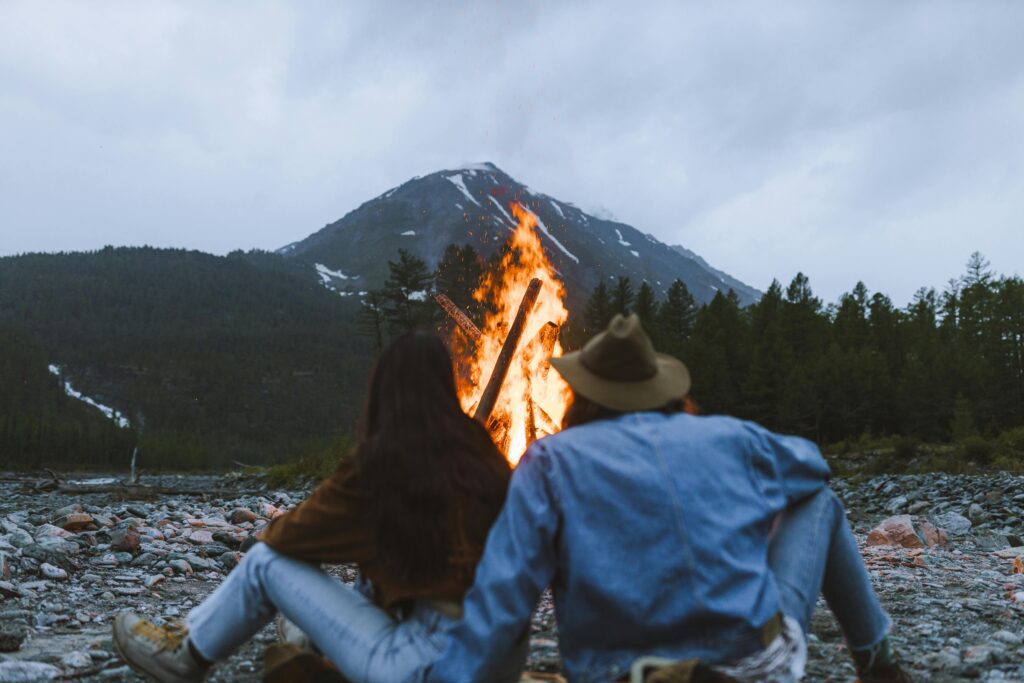 A couple enjoys a warm bonfire in a serene mountain landscape, surrounded by nature.